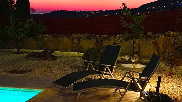 Uma área à beira da piscina com espreguiçadeiras e uma mesa, cercada por um muro de pedra e árvores.