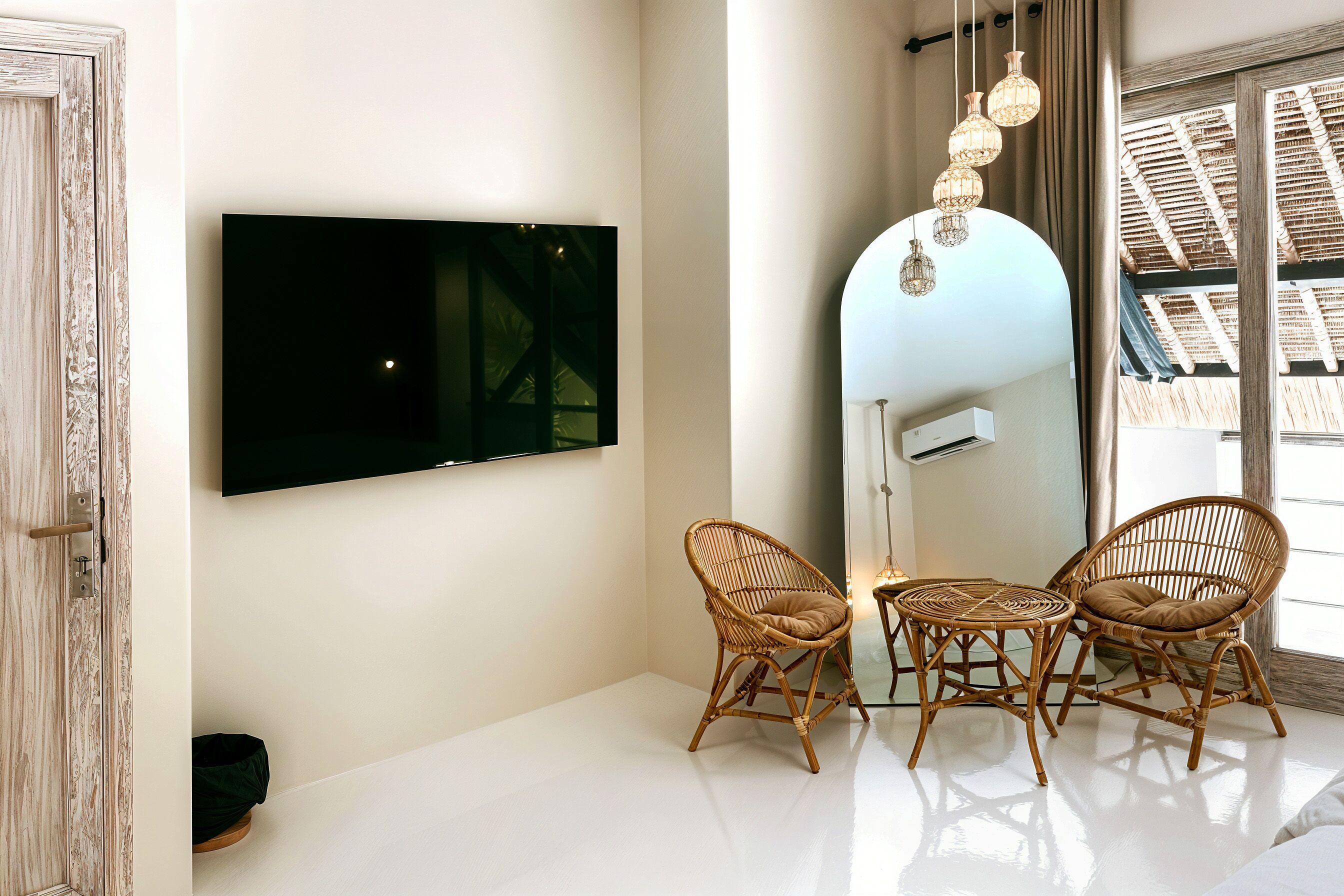 Een moderne kamer met een flatscreen televisie aan de muur, twee rieten stoelen, een kleine ronde tafel en een grote spiegel die het plafond reflecteert.