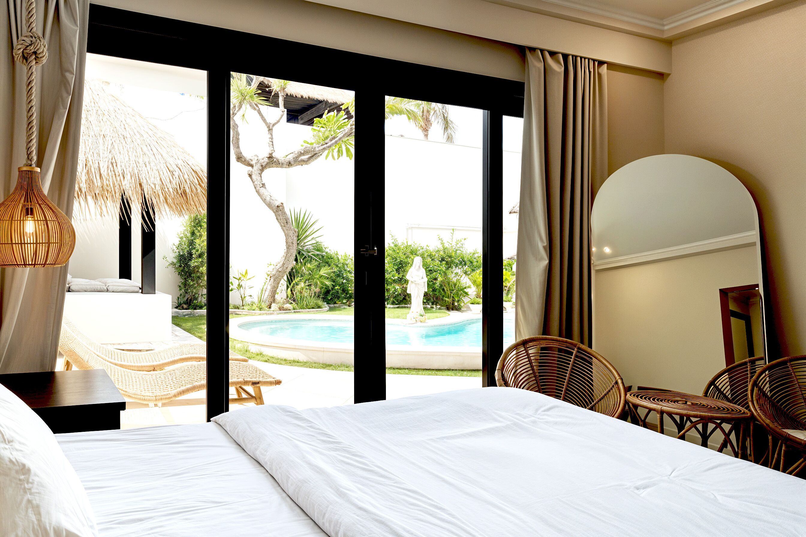 Camera d'albergo con un letto, sedie in vimini, un tavolino, uno specchio grande e vista su una piscina e vegetazione.