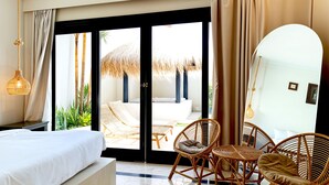 A hotel room with a bed, wicker chairs, a dining table, and a view of an outdoor seating area with a thatched roof. - Villa de Malicott (Seminyak)