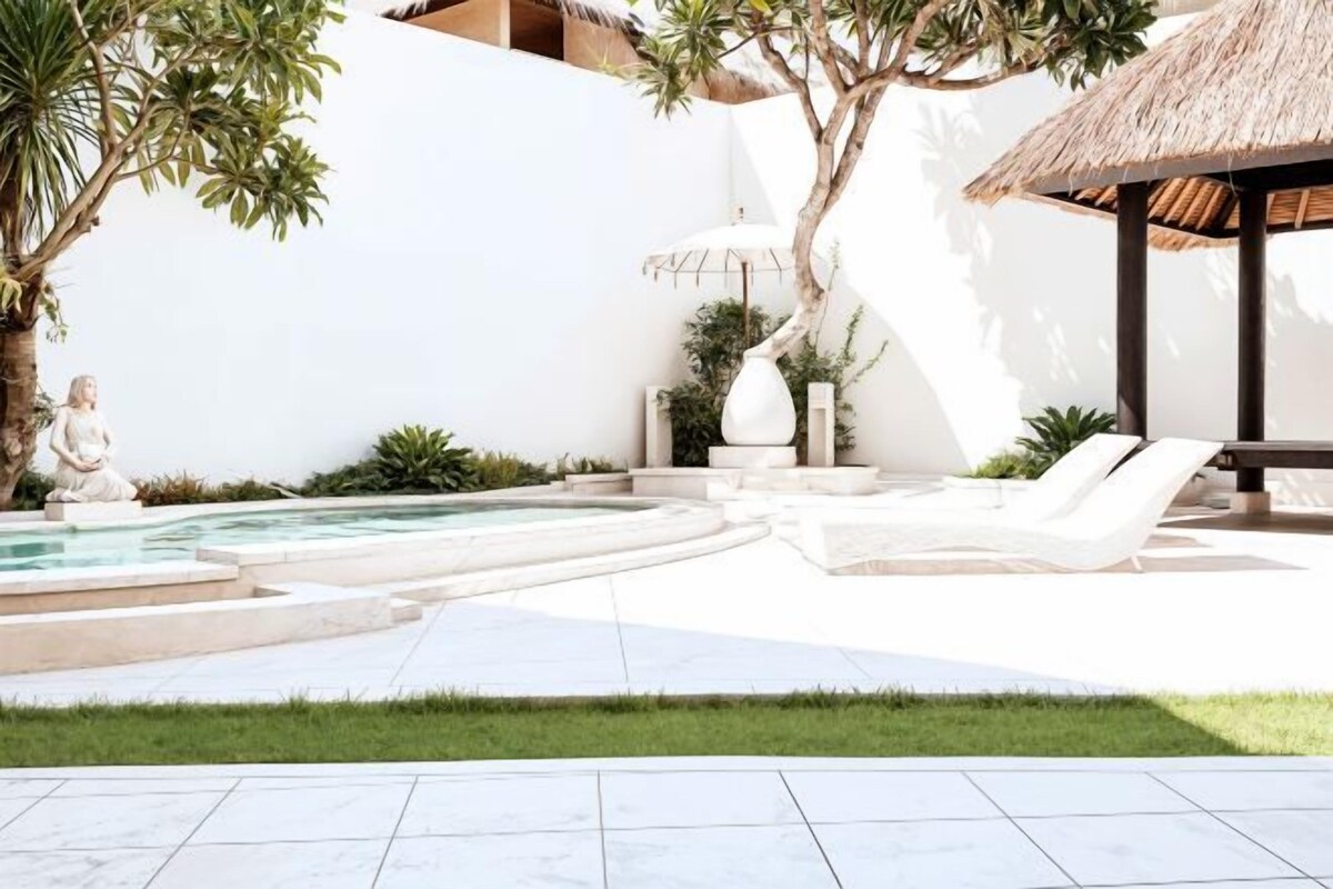 A modern outdoor pool area with lounge chairs, a statue, and a thatched-roof structure.
