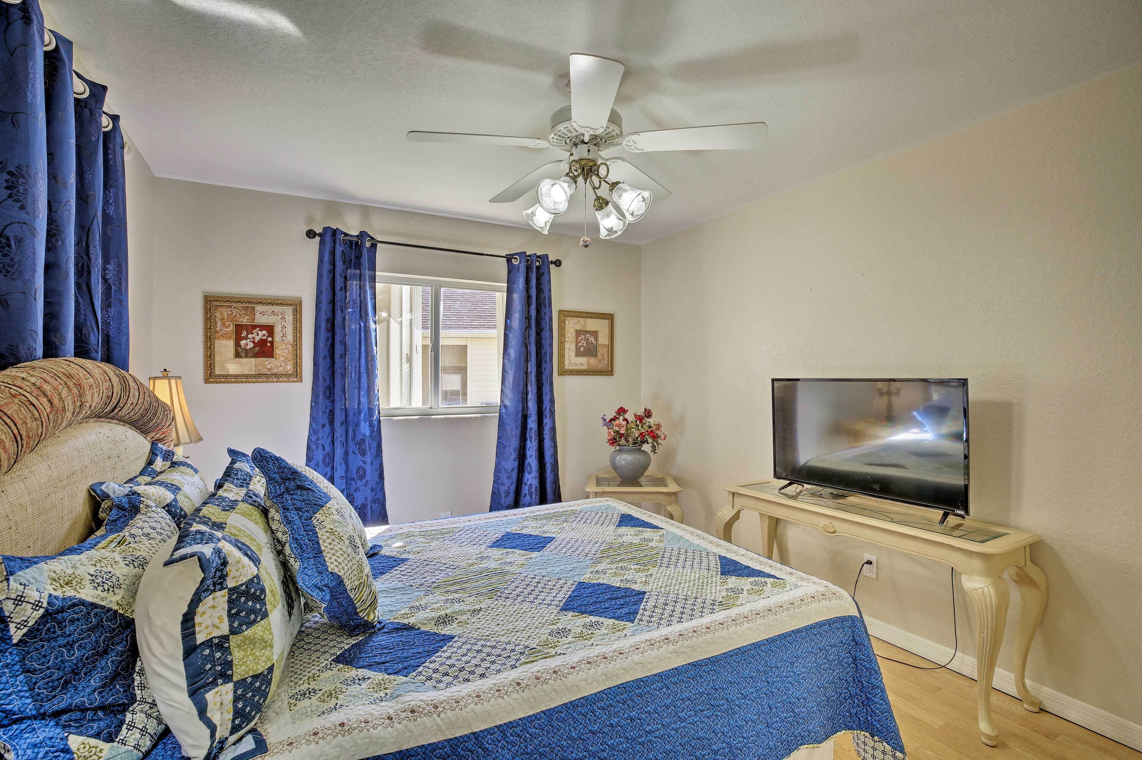 A bedroom with a bed, a television on a stand, a ceiling fan, and curtains.
