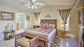 Ein Schlafzimmer mit einem Bett, einem Holzsessel, einem Nachttisch, einem Deckenventilator und einem Fenster mit Vorhängen.