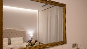 A bedroom with a large ornate mirror, a bed with white linens, a nightstand with a lamp, and a vase with flowers. - B4B Sea n' City (Zakynthos)