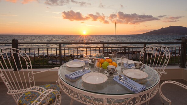 A round glass table set for dining with chairs, overlooking a sunset over the sea. - B4B Sea n' City (Zakynthos)