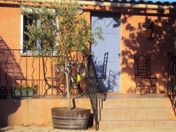 A building with a blue door, a small tree in a barrel planter, and two metal chairs on the porch. - Studio La Douloire (Pernes-les-Fontaines)