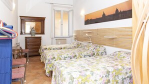 Una habitación con dos camas, cabecera de madera, ventilador de techo, ventana con persianas, radiador, armario de madera y un cuadro de un paisaje urbano.