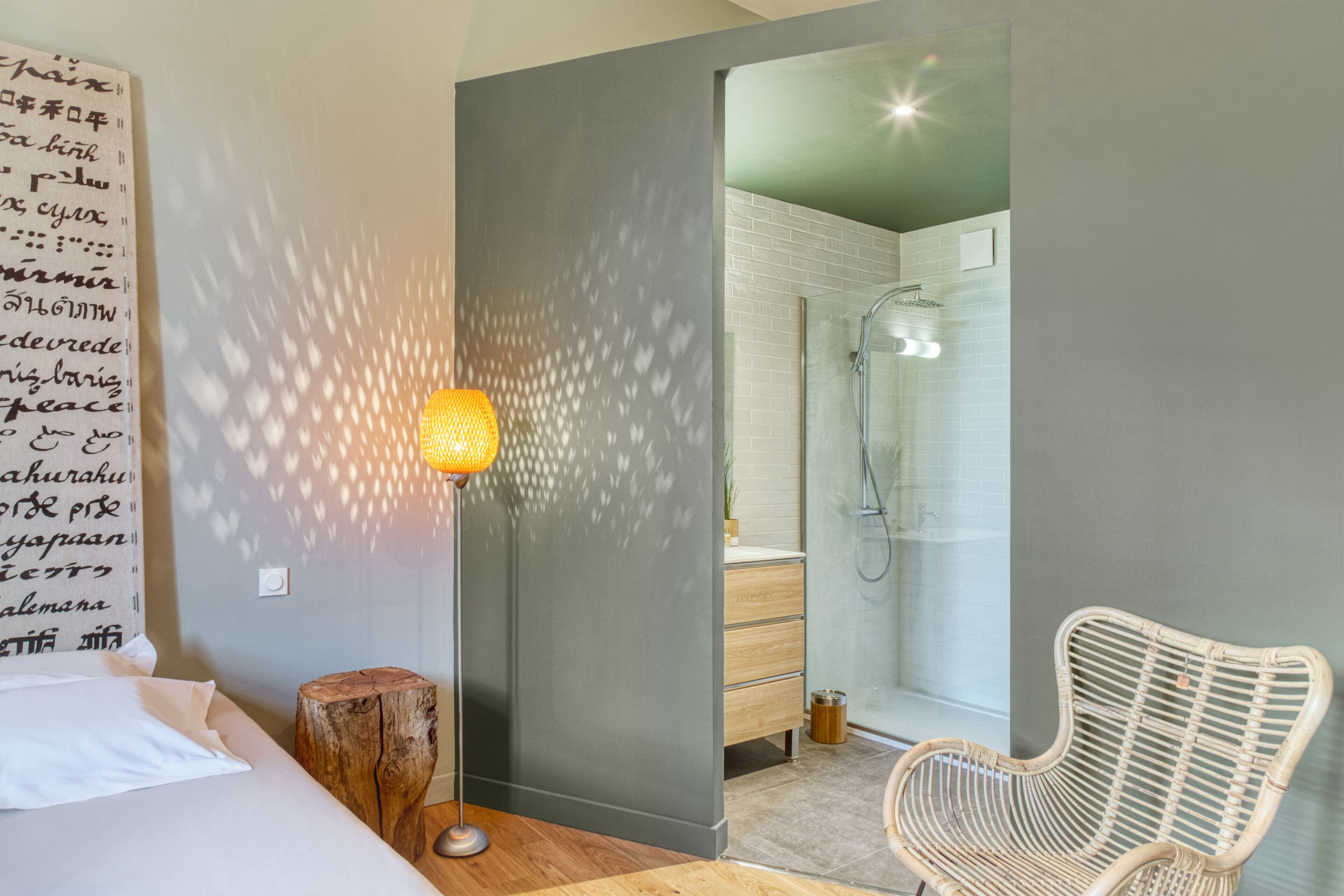 A modern hotel room with a bed, a standing lamp, a wicker chair, and a bathroom with a glass shower.