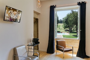 Een kamer met een groot raam, een tafel met een koffieapparaat, twee stoelen en een schilderij aan de muur.