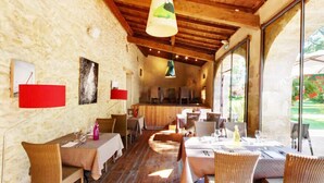 Un espace de restauration rustique avec des poutres en bois, des murs en pierre et de grandes fenêtres. Des tables sont disposées avec des chaises, et une scène est visible au fond.