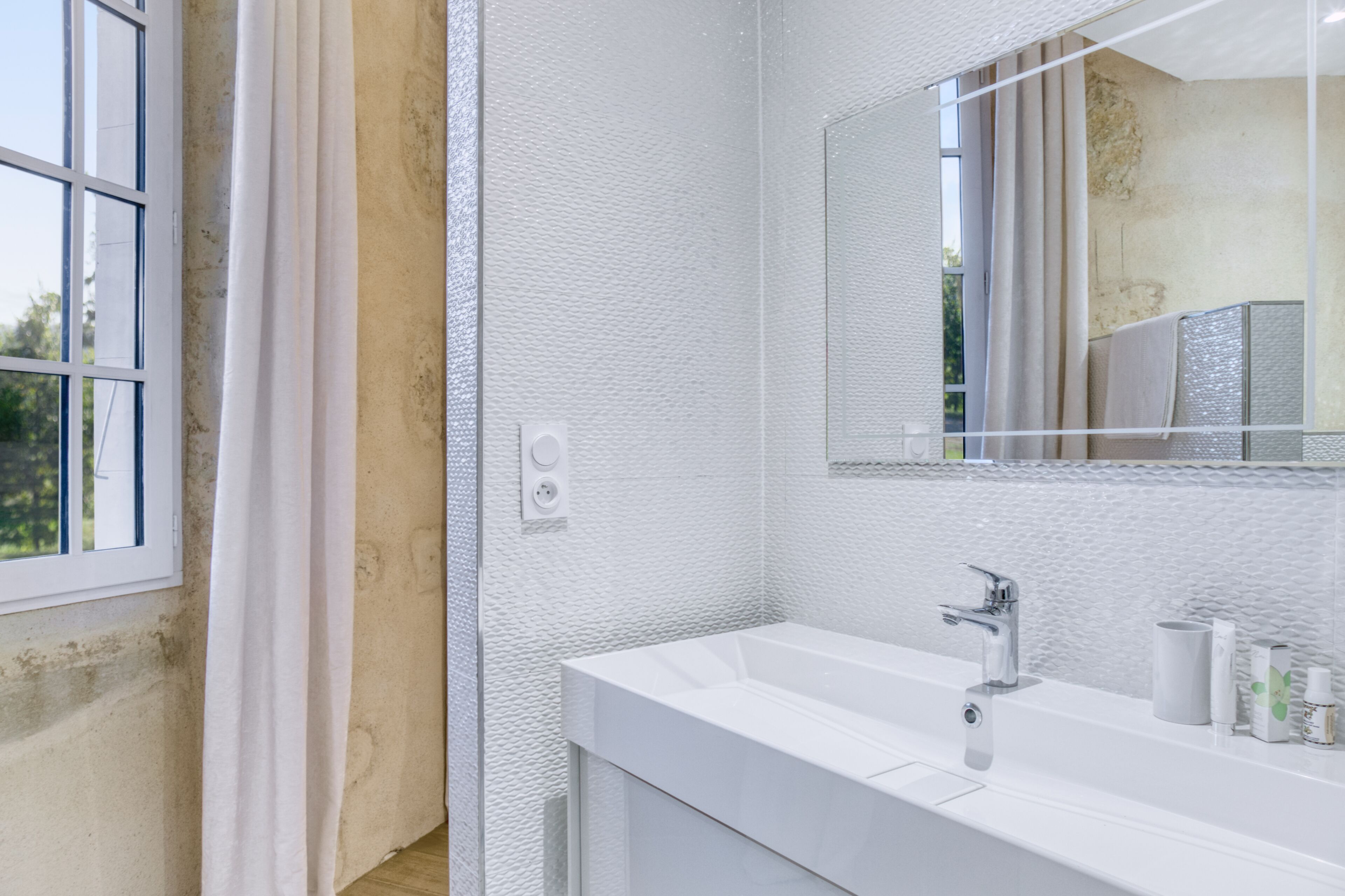 Een moderne badkamer met een wit wastafelmeubel, een grote spiegel en een raam met gordijnen.