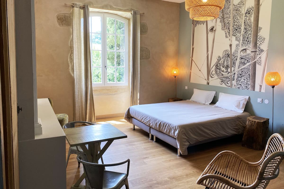 Un dormitorio con cama, escritorio, dos sillas, ventana con cortinas, una lámpara de pared y un tapiz.