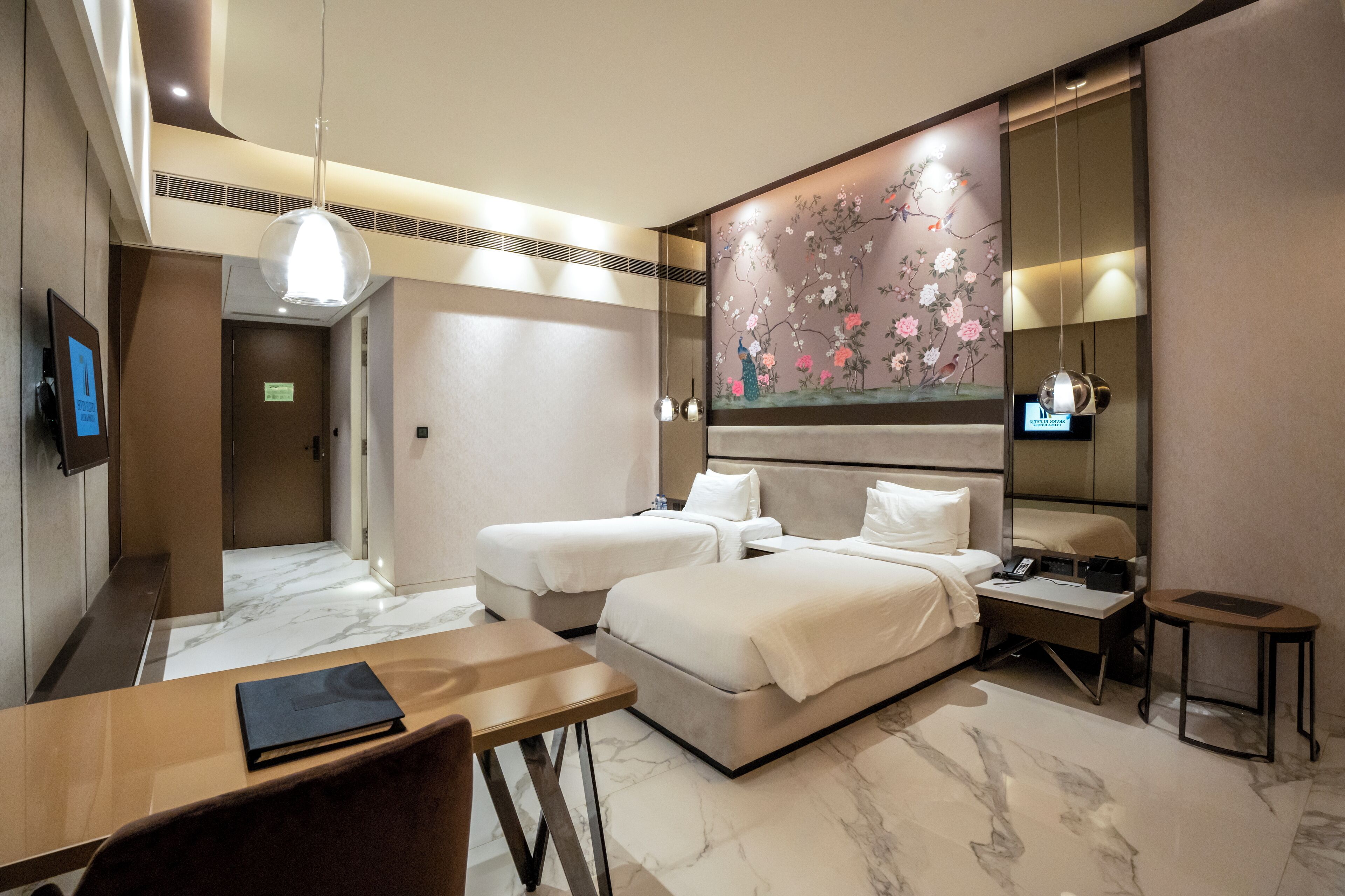 A hotel room with two beds, a desk, a chair, a TV, and a large floral artwork on the wall.