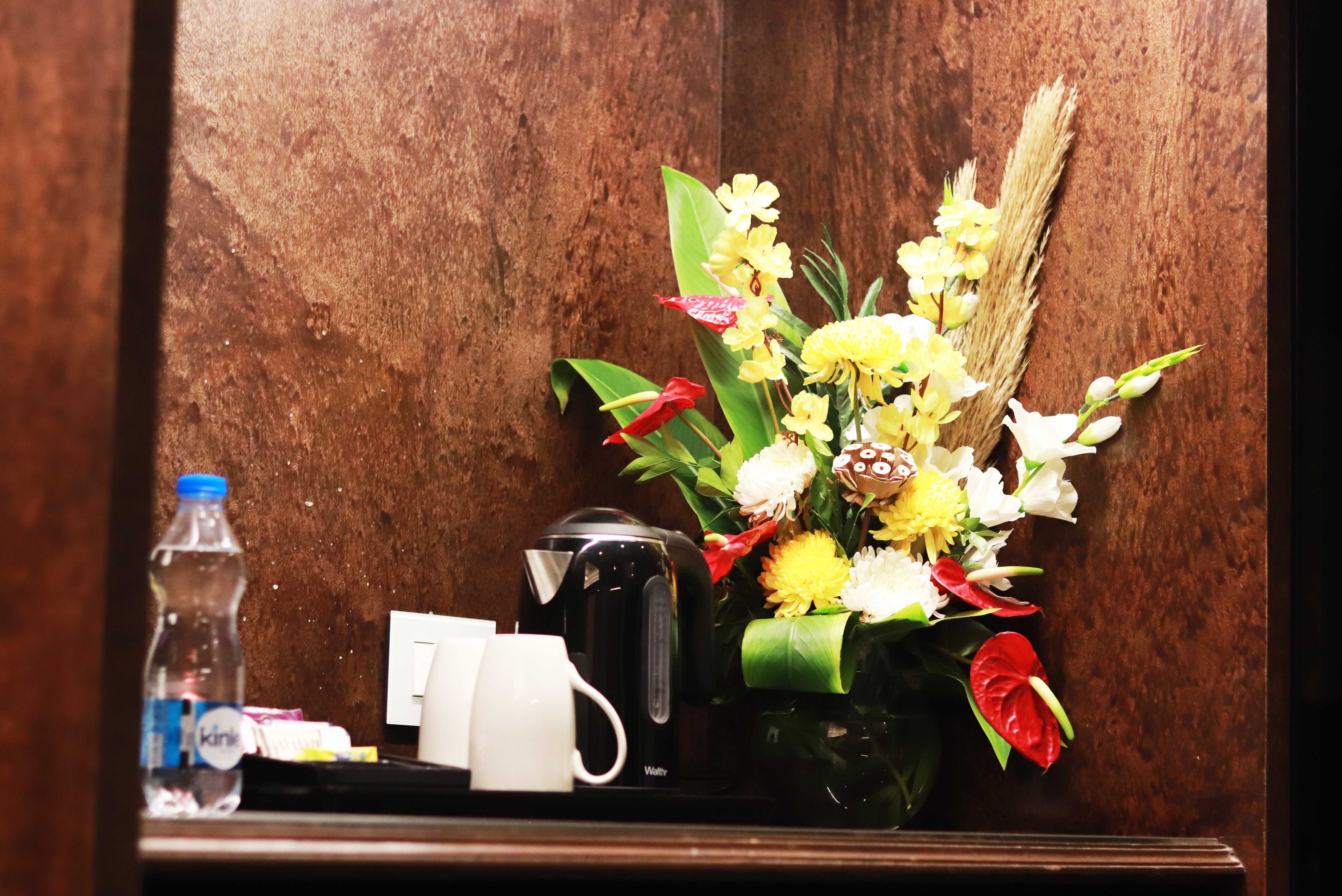 A wooden wall with a small table holding a water bottle, a white mug, a black kettle, and a vase with flowers.
