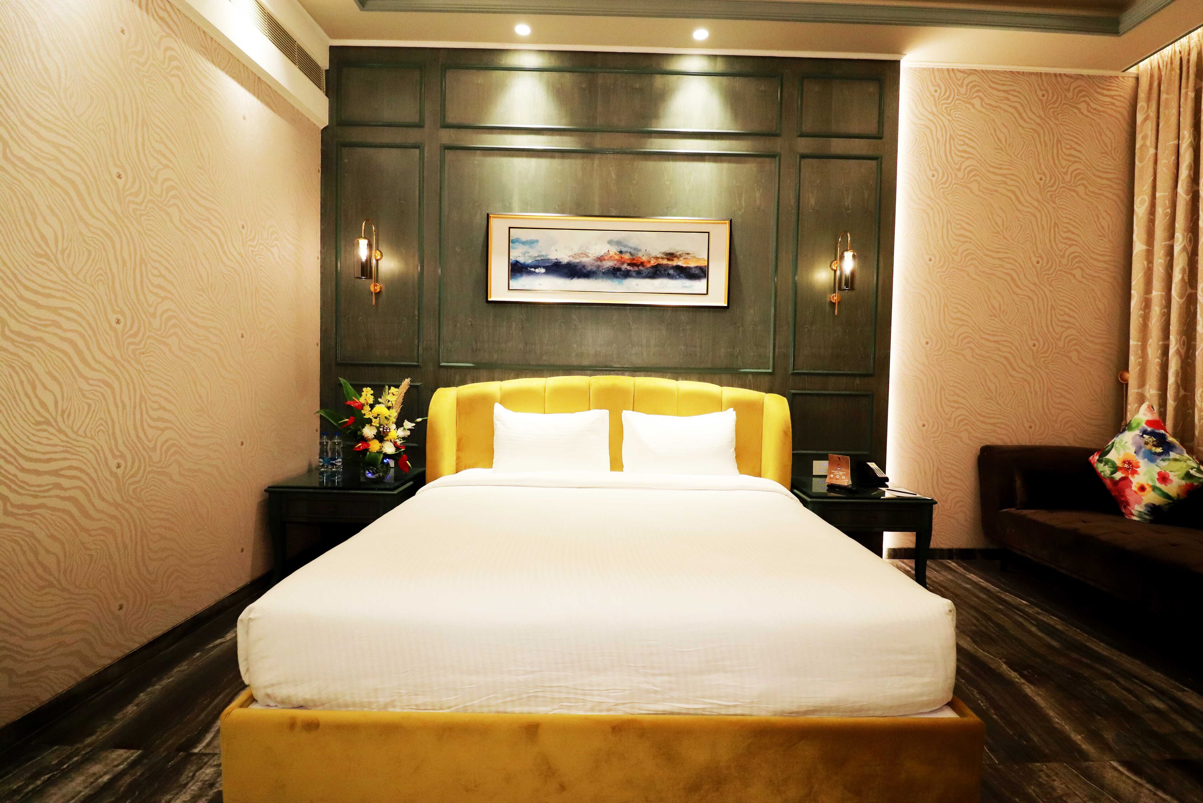 Habitación de hotel con una cama grande, una mesita de noche con un florero y un cuadro en la pared, además de un piso de madera oscura.