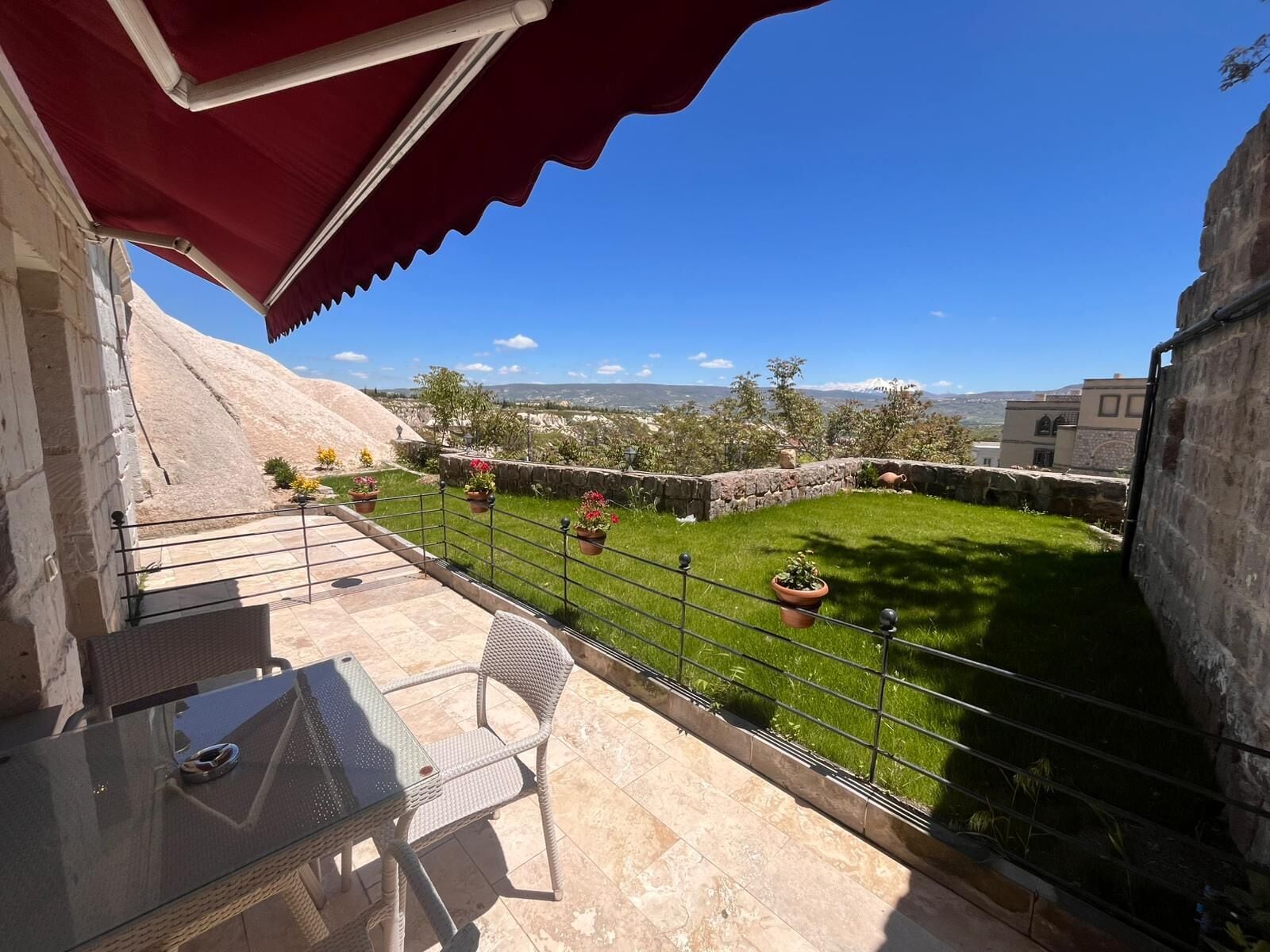 A patio with a glass table and wicker chairs, a red awning, and a view of a grassy area with potted plants.