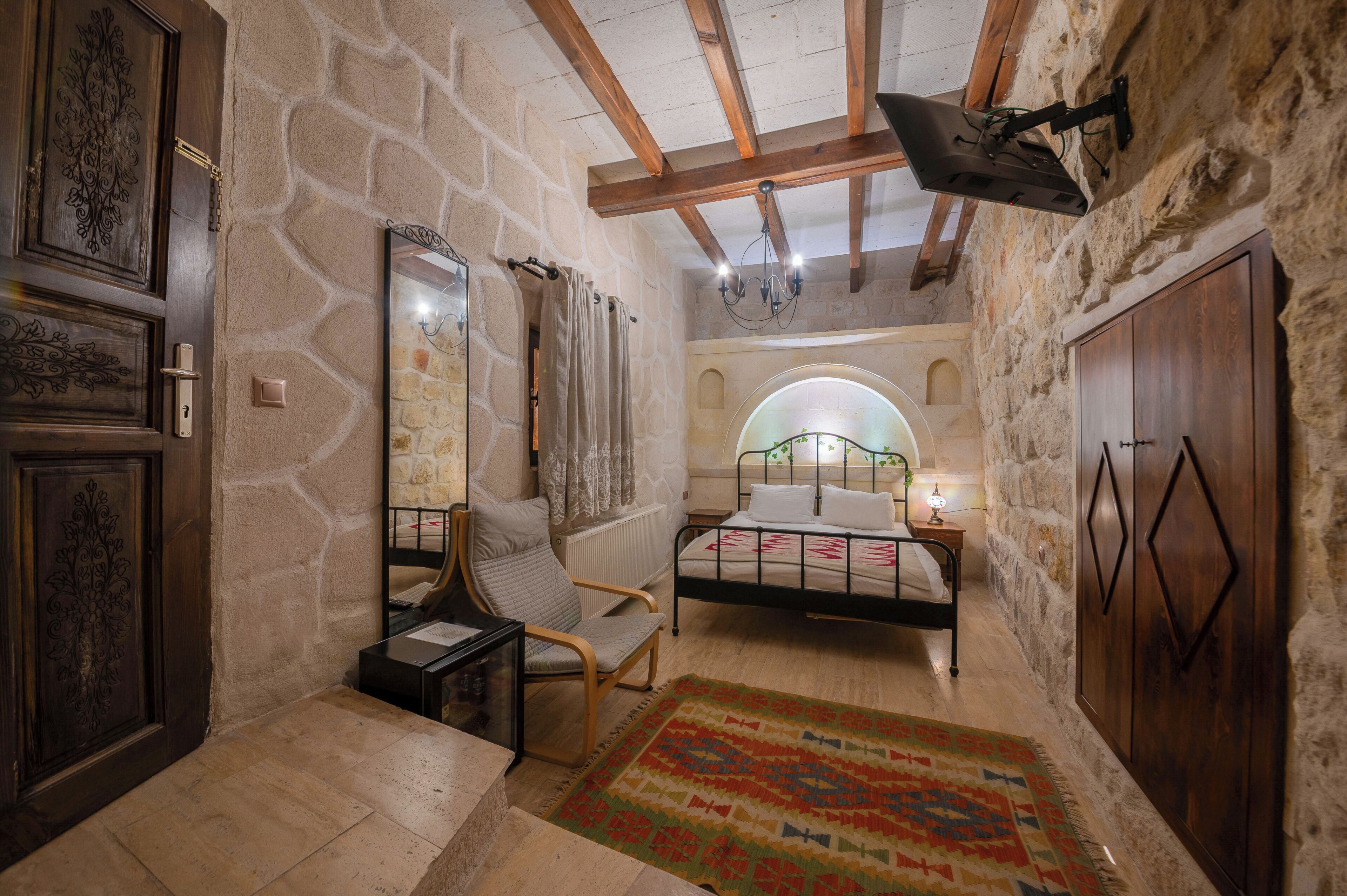A bedroom with stone walls, a wooden ceiling, a bed, a chair, a mirror, and a television.