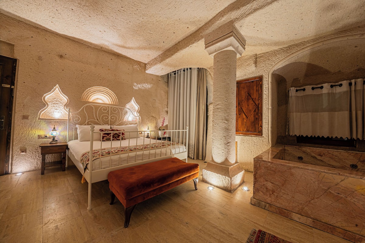 Quarto com cama, banqueta, mesa de cabeceira, abajur e banheira.