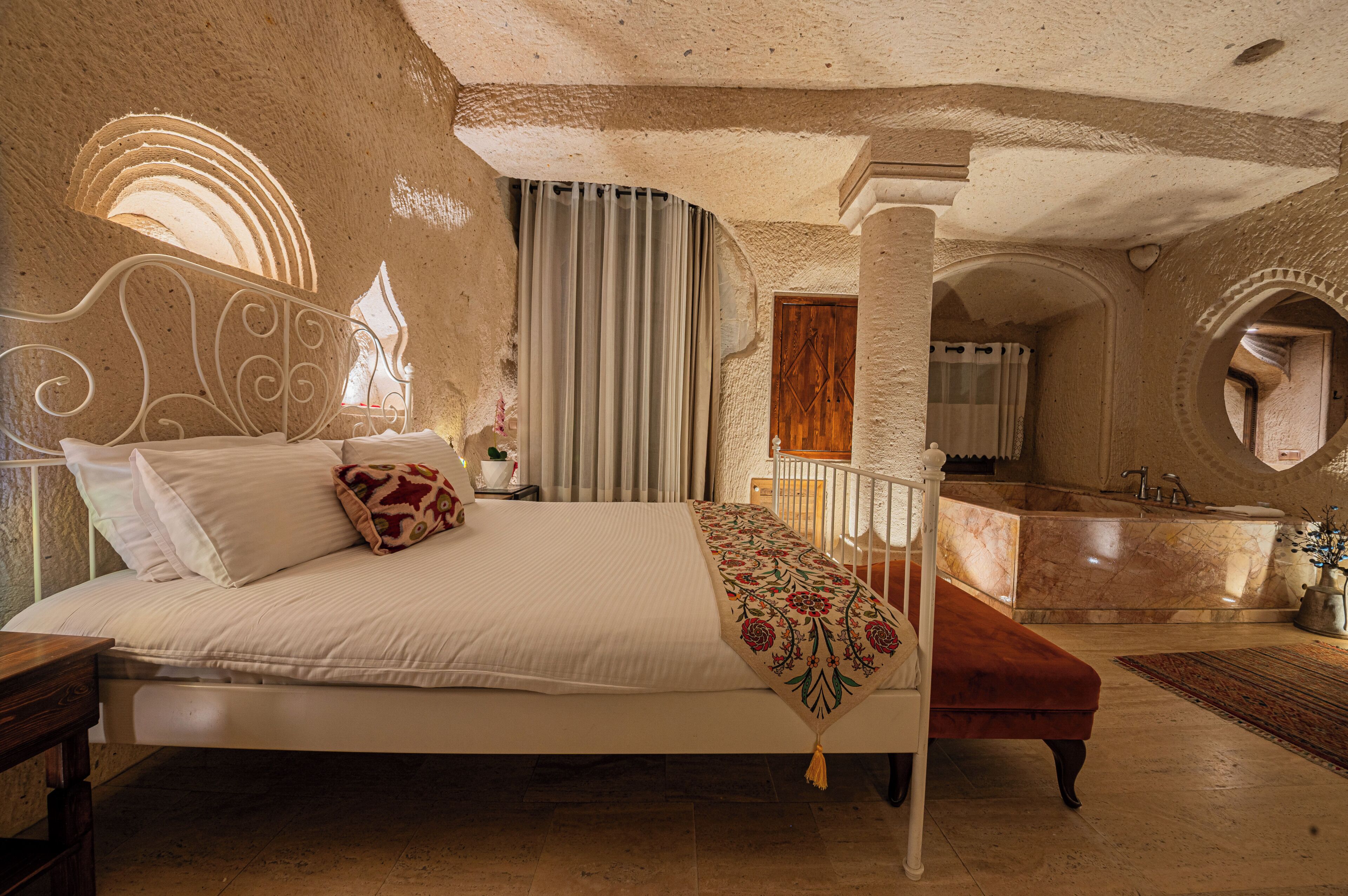 Un dormitorio con cama, mesita de noche, ventana con cortinas, baño con bañera y espejo.