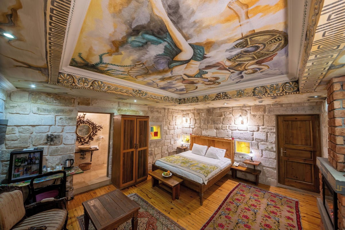 Quarto com parede de pedra, cama com cabeceira de madeira, um banco, uma mesa, um espelho e lareira.