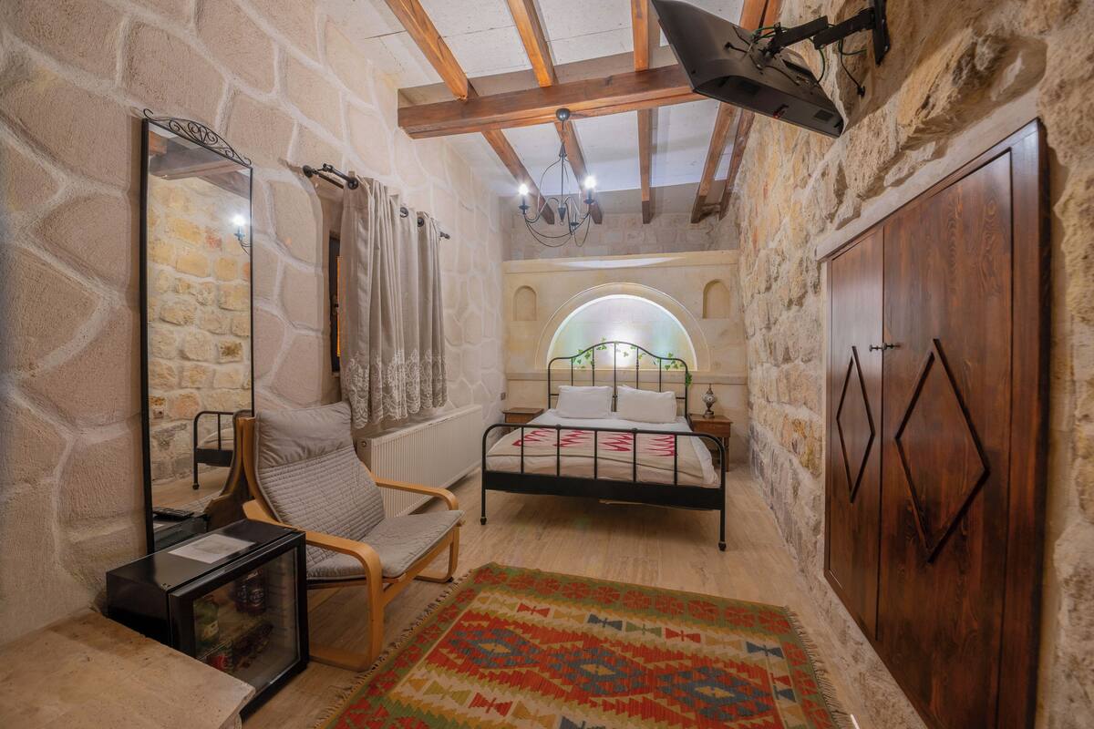 Quarto com paredes de pedra, teto de madeira, cama, cadeira, guarda-roupa e espelho.