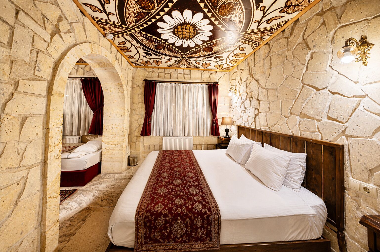 A room with a large bed, a wooden headboard, stone walls, a decorative ceiling, and a window with curtains.