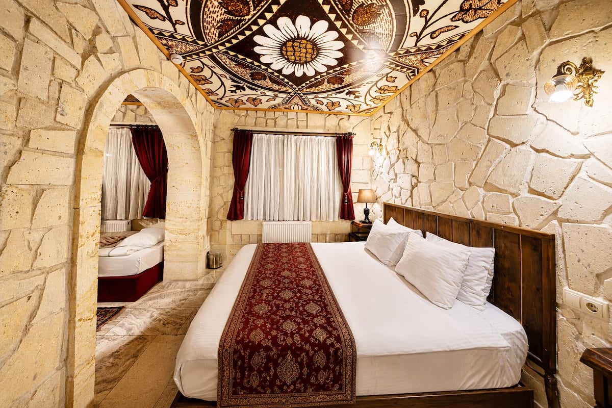 Quarto com uma cama grande, cabeceira de madeira, paredes de pedra, teto decorativo e janela com cortinas.