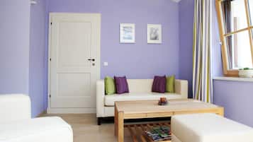A room with a white door, a white sofa with green and purple cushions, a wooden coffee table, and a window with striped curtains.