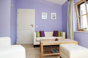 A room with a white door, a white sofa with green and purple cushions, a wooden coffee table, and a window with striped curtains.