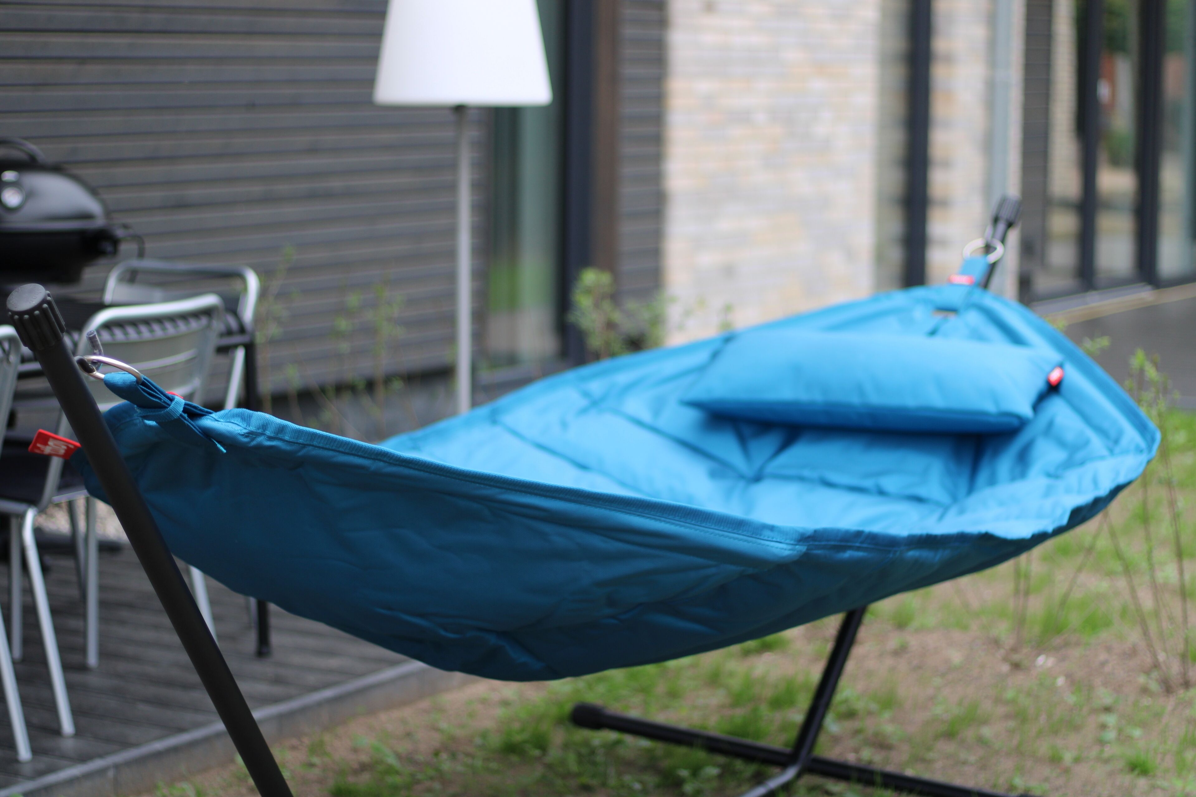 Een blauwe hangstoel buiten, met een metalen standaard, een tafel en stoelen op de achtergrond.