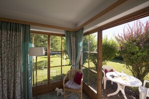 A sunroom with a view of a garden, a wicker chair, a small table, and a lamp. - Gutshof Wolfgangsee Resort & Event Hotel (Strobl)