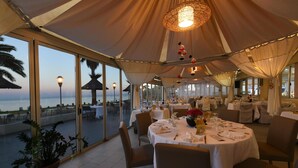 An indoor dining area with tables set for guests, large windows offering a view of the sea, and a tented roof structure. - Residence San Pellegrino (Penta-di-Casinca)