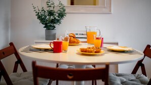 Una mesa redonda de comedor con alimentos para el desayuno, como cruasanes, jugo de naranja y una taza roja. Una planta en maceta está en la pared detrás de la mesa.