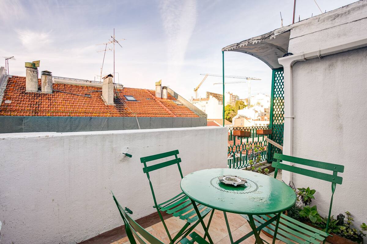 Eine Dachterrasse mit einem grünen Tisch und Stühlen, einer weißen Wand und Blick auf Gebäude mit roten Ziegeldächern.
