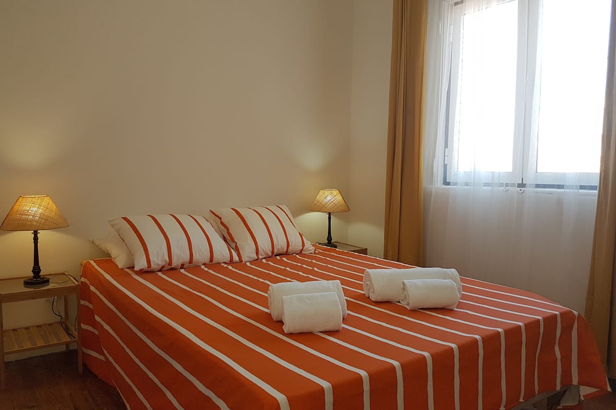 A hotel room with a bed, two bedside lamps, a window with curtains, and a wooden nightstand.