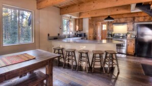 A rustic kitchen with wooden beams, a large island with stools, and a view of the forest through the window. - Enchanting Chamberlands Cabin (Tahoma)