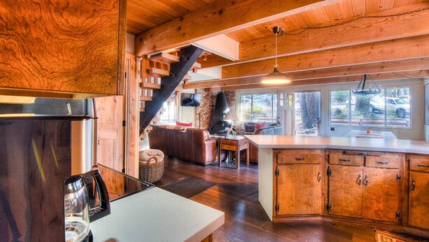 A kitchen with wooden cabinets, a stainless steel refrigerator, a dining area with a table and chairs, and a large window offering a view of the outdoors. - Enchanting Chamberlands Cabin (Tahoma)