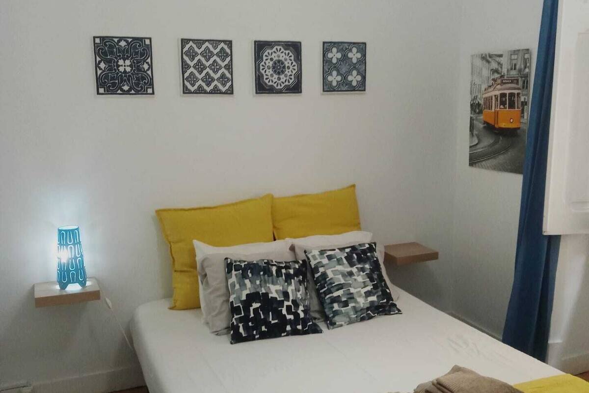 A bedroom with a bed, pillows, a nightstand, a wall-mounted lamp, and a picture of a tram.