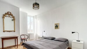 A bedroom with a wooden floor, a bed with grey bedding, a desk, a chair, a bedside table with a lamp, a mirror, and a window with natural light.