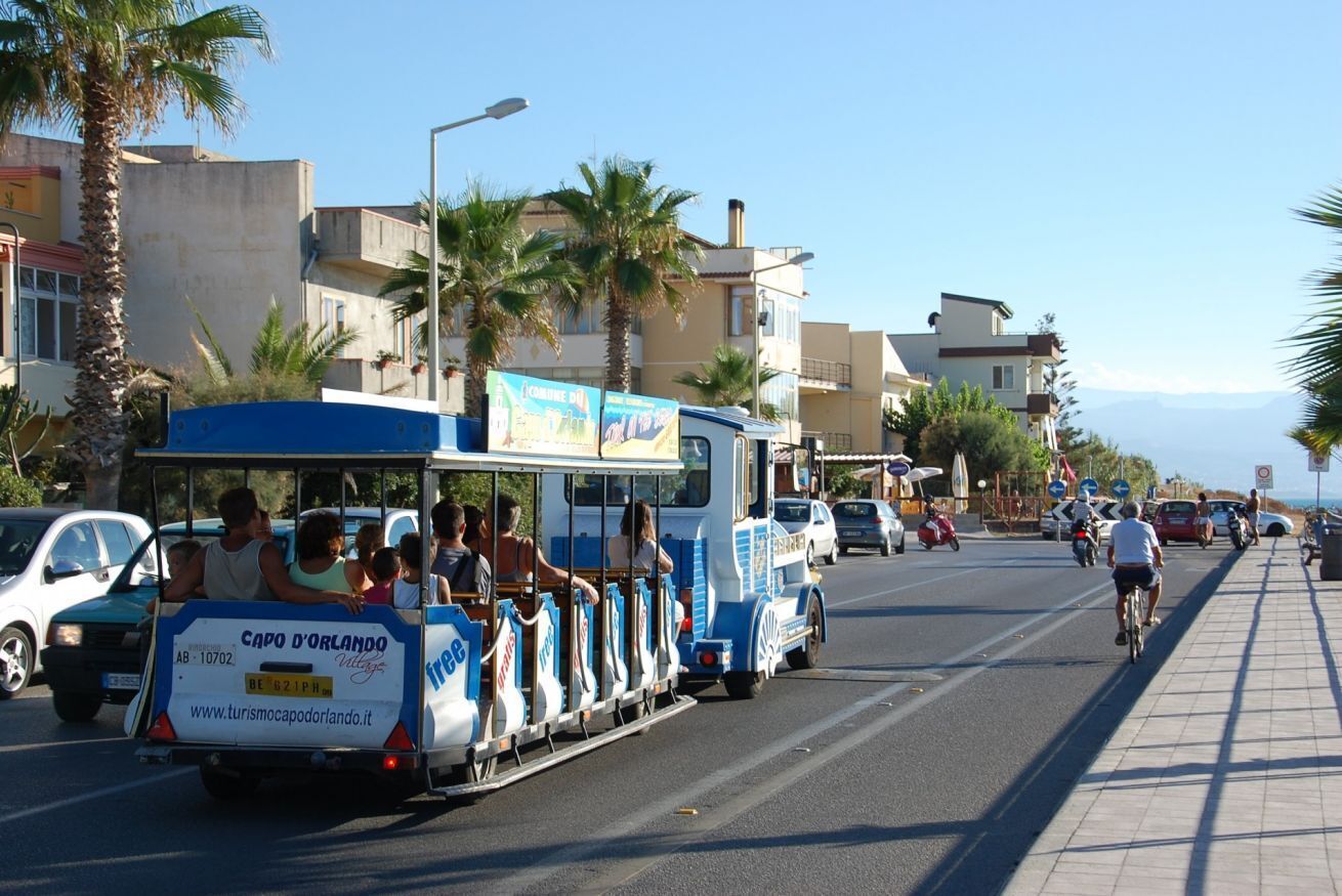 Un tram touristique rempli de passagers est garé dans une rue, avec des bâtiments et des palmiers en arrière-plan.