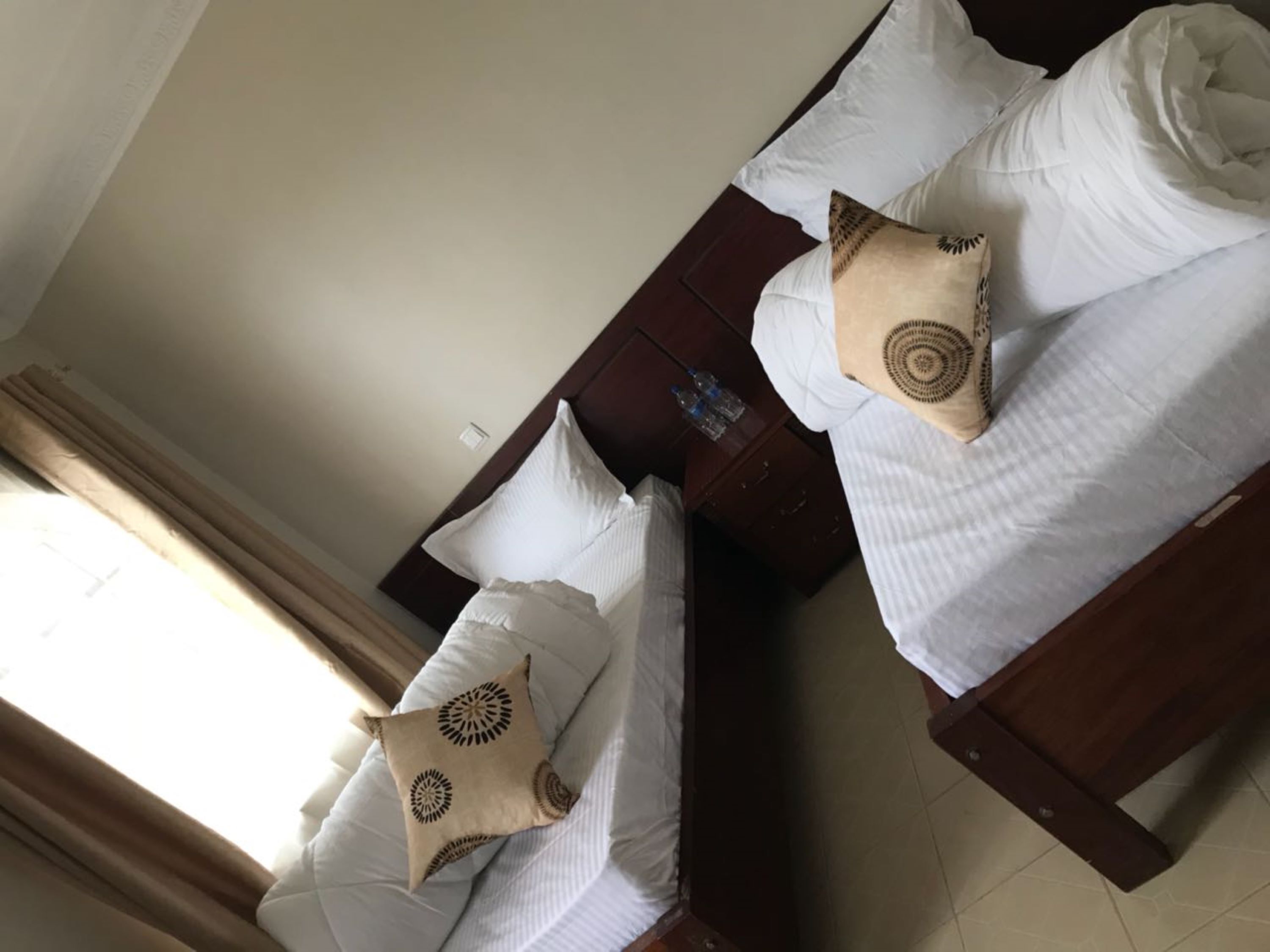Dos camas con sábanas blancas y almohadones decorativos, un cabecero de madera, una mesita con una botella de agua y una ventana con cortinas.