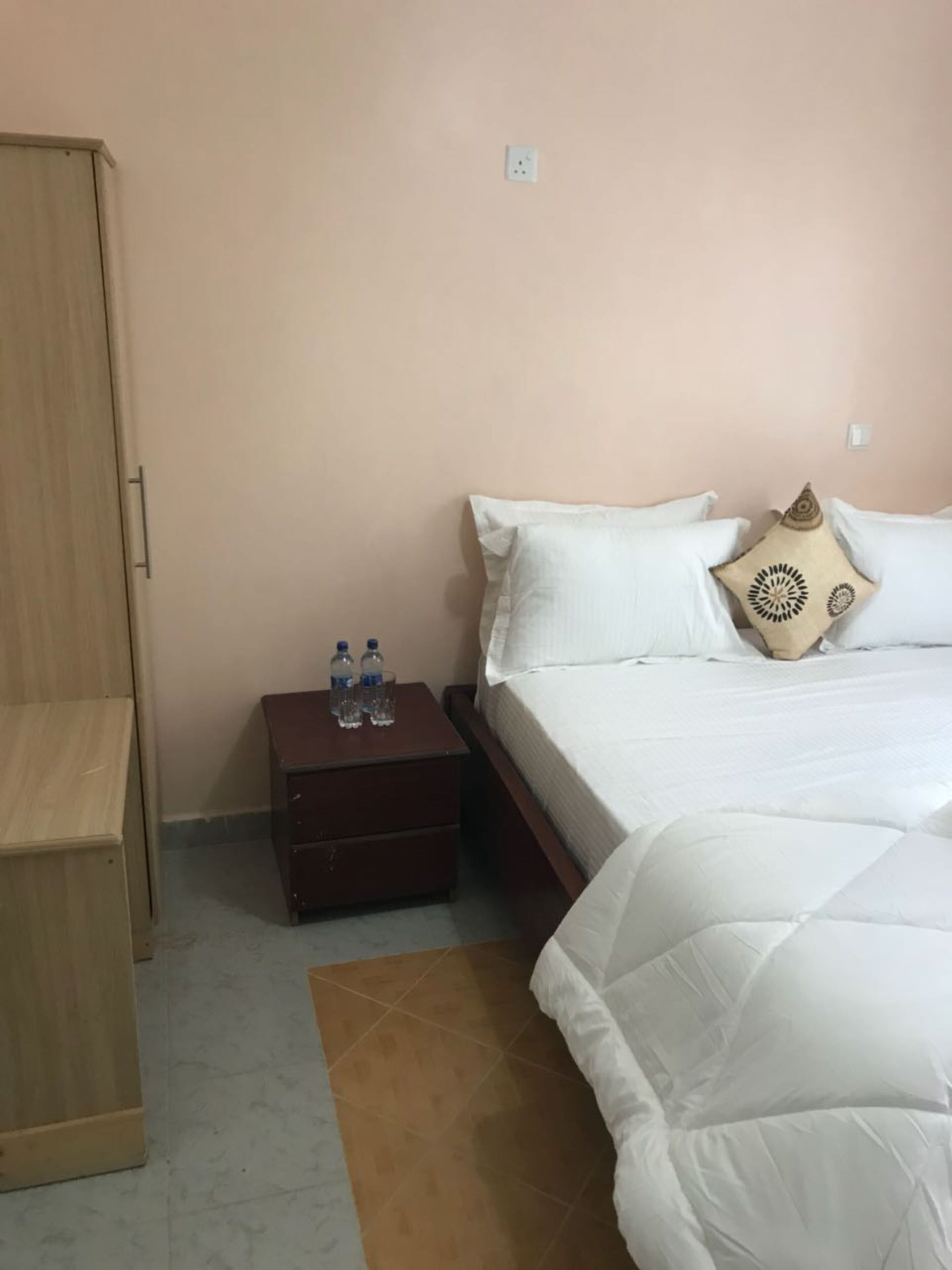 A bedroom with a bed, a nightstand with water bottles, a wardrobe, and a brown rug.