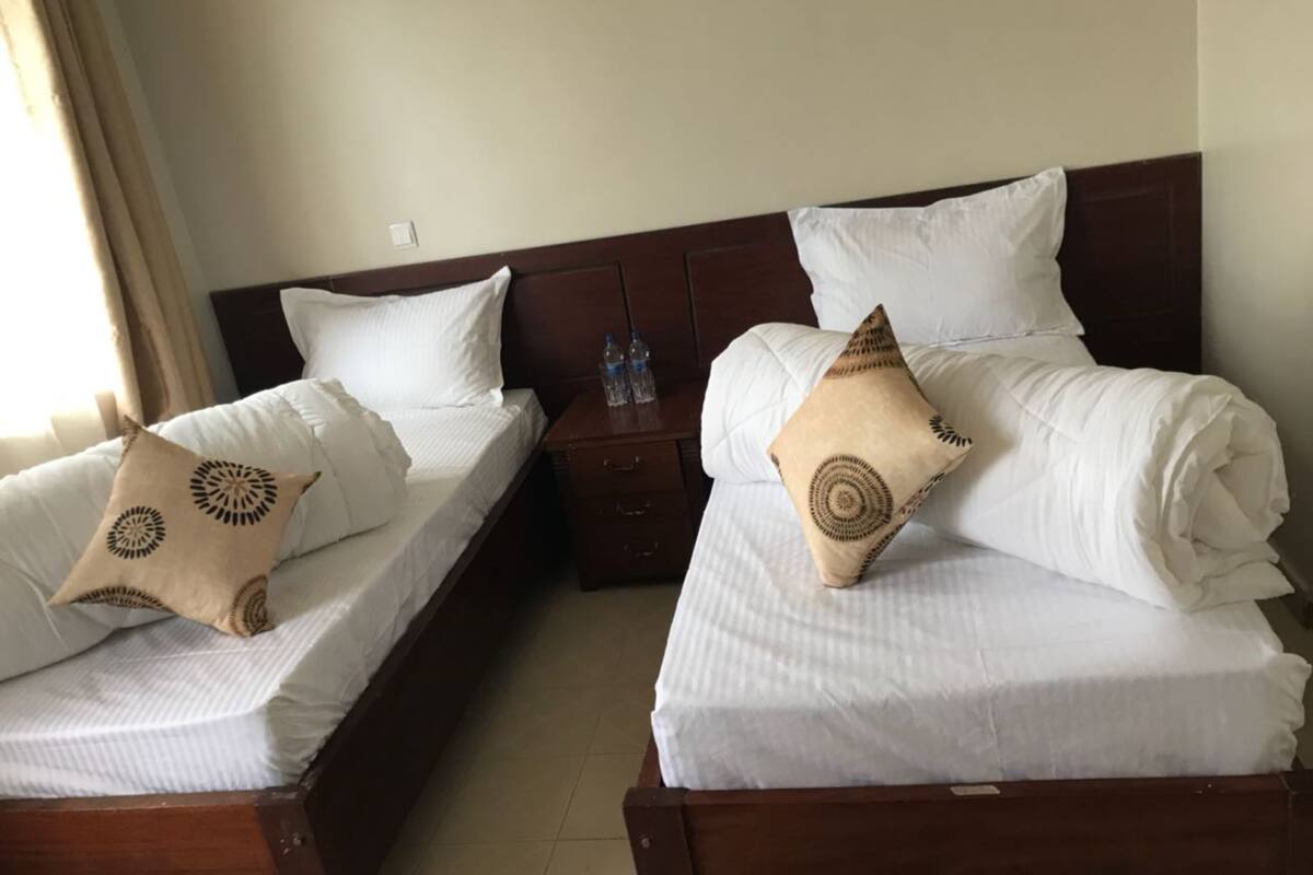 Two single beds with white linens and decorative pillows, a wooden headboard, a nightstand with water bottles, and a window with curtains.