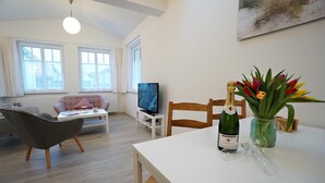 A modern living room with a sofa, a television, a dining table with a bottle of champagne and glasses, and a vase of flowers.