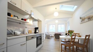 A modern kitchen with white cabinetry, a built-in oven, and a coffee maker. There is a dining area with wooden chairs and a table, and a skylight above.