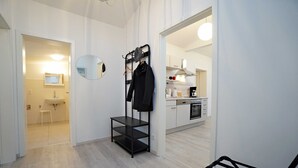 A modern hotel room with a wooden floor, a black metal rack with clothes, a round mirror, and a view into the kitchen area.