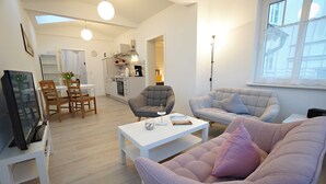 A modern living room with a sofa, armchair, and coffee table. There is a TV on a stand and a painting on the wall.