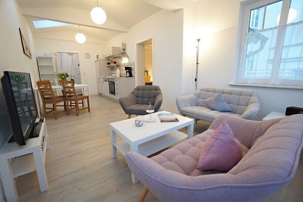 A modern living room with a sofa, armchair, and coffee table. There is a TV on a stand and a painting on the wall.