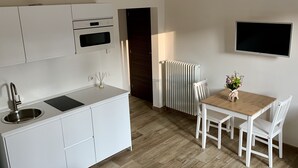 Una cocina moderna con armarios blancos, horno empotrado y un lavabo. Hay un comedor con una mesa de madera y sillas blancas, y un radiador en la pared.
