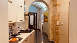 A narrow kitchen with a gas stove, cabinets, and a tiled floor. An archway leads to a hallway with a painting on the wall.